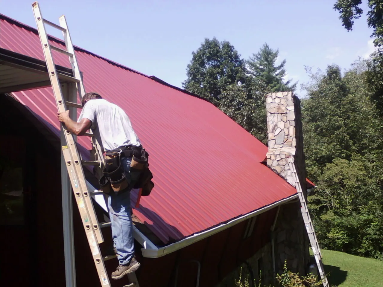 Roofer on a ladder beside a red metal roof and stone chimney — Soffit & Fascia Repair by Roofing Brothers in Buffalo Lake